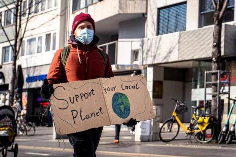 man wearing black gloves, orange jacket, mask, and a red knit hat; he's holding a sign that says "support your local planet"