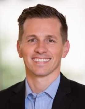 A White man with brown hair smiling wearing a blue collar shirt and black blazer.