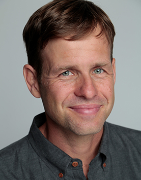 A White man smiling with brown hair wearing a grey collar shirt