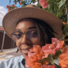  A photo of Zel wearing a denim shirt, a straw hat over locs and wood-framed glasses while smiling behind pink bougainvillea flowers.