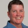 A white women smiling with a short pixie cut wearing a light blue collared shirt. 