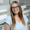 Latina women with light caramel skin, light brown long straight hair. She is wearing blue square glasses and a light blue blouse. She is looking to the side and standing in front of a glass wall with horizontal white and light blue lines.