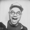A classic black and white photo booth portrait of a man wearing circular glasses and a trucker hat with an exaggerated smile.