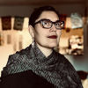 Female with purple frame glasses, black hair pulled back, black with white specs shawl. Background is colorful with decorative Mexican paper.