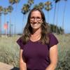 women with shoulder length brown hair and glasses in a maroon shirt. in the background are palm trees and a blue sky