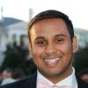 Indian American male wearing suit with bowtie smiling. A white State house blurred in the background