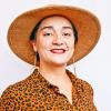 Latinx/Chicana female with light olive tone skin wearing red lipstick standing against a white backdrop. Person is wearing a brown/rust colored wool hat and a black and brown leopard button down shirt.
