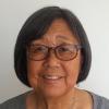 Kathy is an Asian American/Japanese American woman with chin-length dark brown hair with some gray.  She is smiling and is wearing tortoise shell glasses and silver earrings. She has on a gray scoop-necked top and is framed against a white background.