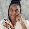Brown skin Black woman with dreadlocks holding her hand to the side of her face in a contemplating pose. She is wearing a white shirt with black, yellow and orange flower graphics.