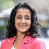 Rochelle is a Sri Lankan woman, smiling while posing in a pink blazer with courtyard umbrellas and green shrubs behind her.