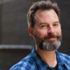 A smiling man with short hair and a dark and light beard, wearing a blue plaid shirt.