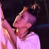 Photograph of a Japanese American person with a shaved head and top bun and in a large flowing cream colored shirt, outside at nighttime, kneeling on the ground, holding with both hands a long thin piece of fabric up into the air, and looking up at the fabric.