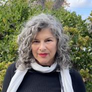 White woman with silver curls wearing a black top and a white scarf.