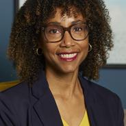 Black, brown skin woman with curly brown hair wearing grey glasses, yellow blouse and blue blazer.