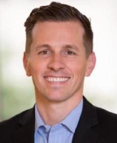 A White man with brown hair smiling wearing a blue collar shirt and black blazer.