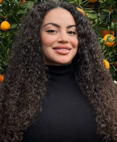 A woman smiling with a black shirt and curly long brown hair in front of an orange tree.