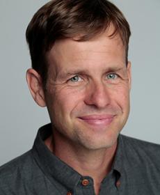 A White man smiling with brown hair wearing a grey collar shirt